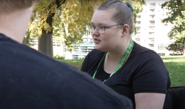 Photo of a white person wearing a sunflower lanyard (symbolising invisible disability), in a park