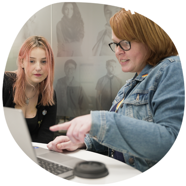 Two white people looking at an open laptop. One of them is pointing at the open screen.