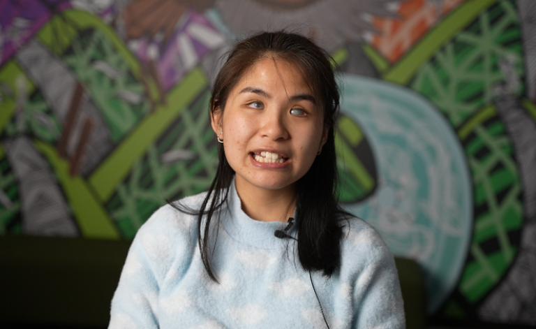 Anna, a CALD young person who is blind, speaking directly to the camera in front of a colourful mural.
