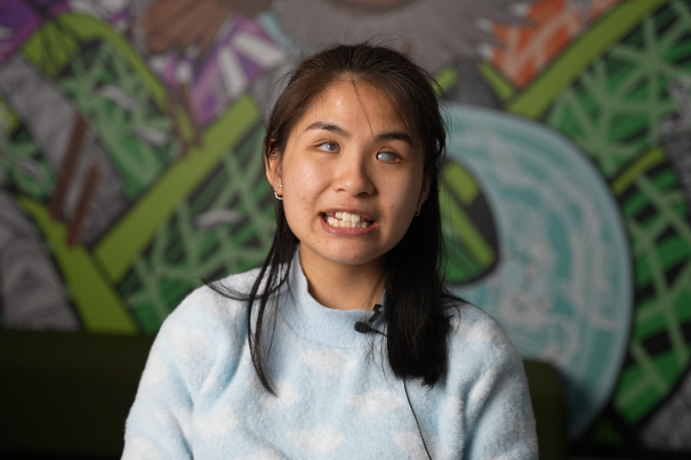 Anna, a CALD young person who is blind, speaking directly to the camera in front of a colourful mural.