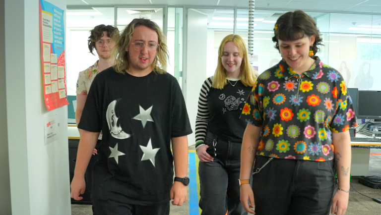 This image shows four young people walking together inside what appears to be an office or community workspace. They are smiling and appear to be enjoying each other's company. The atmosphere seems inclusive and casual, with posters on the wall and computers in the background.
