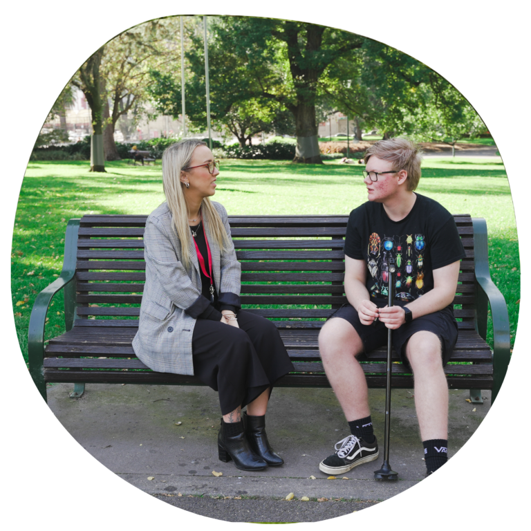 In this photo, two people are sitting on a park bench having a thoughtful conversation. On the left, a woman with long blonde hair and glasses, dressed in a grey blazer and black outfit, faces a young man on the right, who is wearing a black T-shirt with colorful graphics and holding a white cane, indicating he may be blind or have low vision. The setting is a peaceful, green park with trees and sunlight, creating a calm and supportive atmosphere.